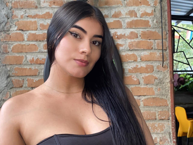 AlaraKey has long black hair, brown eyes, and is wearing a strapless black top, showing her boobs. She's standing against a brick wall, looking confident.