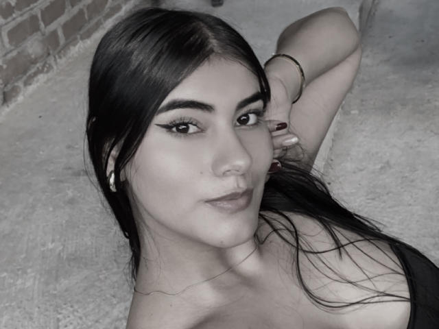 AlaraKey has dark, straight hair, thick eyebrows, and is wearing bold eyeliner. She's lying down, showing off her tits and a gold bracelet. Her nails are painted red.