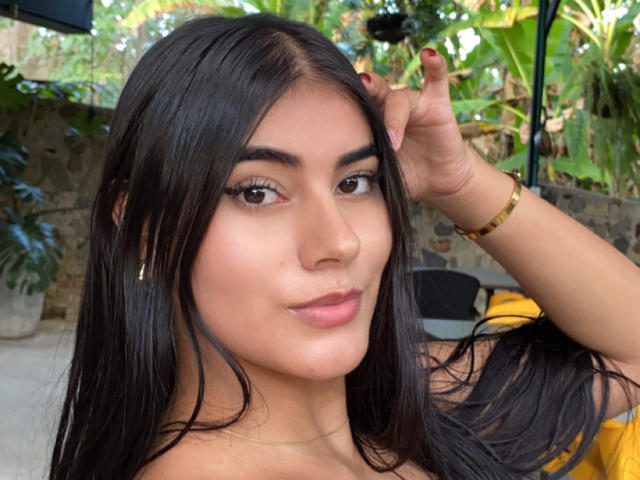 AlaraKey has long black hair, brown eyes, and a slight smile with pink lips. She's touching her hair, wearing a gold bracelet, and has red nail polish.