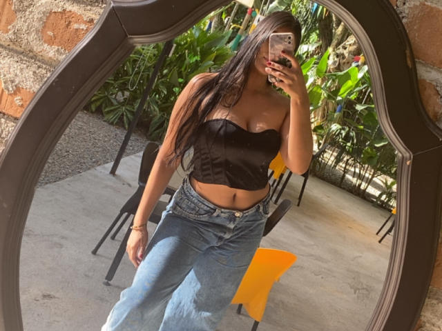 AlaraKey with long brown hair, black strapless top showing her tits, and blue jeans, taking a mirror selfie, showing her bare ass. Her phone has a floral case.