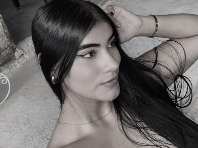 Black-and-white photo of AlaraKey with long dark hair, raised arm, wearing a bracelet, hoop earrings, and eyeliner, looking to the side with a slight smirk. Boobs visible.