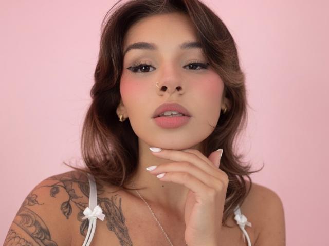 LenaBranch with pink cheeks, brown wavy hair, white floral dress, tattoo on shoulder, gold earrings, hand near chin, pink nail polish, looking at camera, soft pink background.