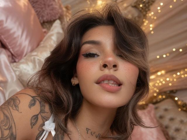 LenaBranch with wavy brown hair, nose ring, and tattoos, has pink lips and eyeliner, looking up with slightly parted pink lips, in a soft pink and gold setting.