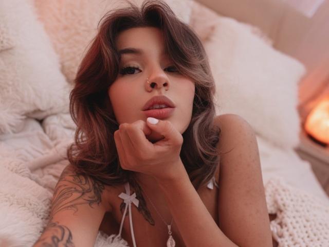 LenaBranch with wavy brown hair, nose ring, and white nail, resting her chin on her hand, showing tattoos on her left arm, lying on white fluffy pillows.