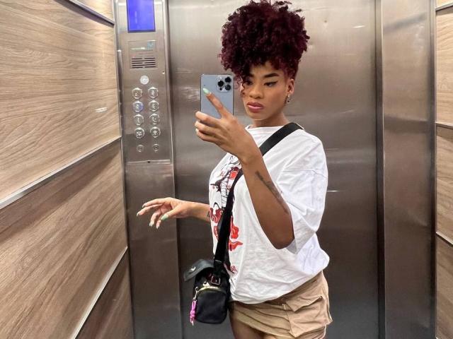 KarlaRicochet, met krullend rood haar, draagt een witte T-shirt en beige shorts, houdt een telefoon vast en staat in een metalen lift. Ze draagt een zwarte crossbodytas en kijkt naar de camera..