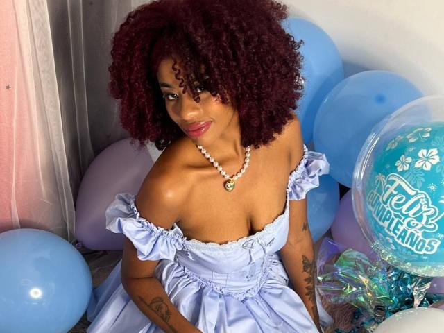Karla, con i capelli rossi ricci, la pelle scura, indossa un abito leggero blu con una spalla scoperta, una collana e siede tra palloncini blu, mostrando tatuaggi sulle braccia, sorridendo. Il seno è visibile..