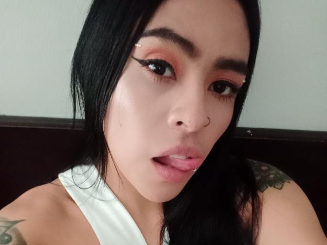StellaBeckker ha i capelli neri, un piercing al naso e del trucco bianco sulle palpebre. Ha la lingua rosa fuori, mostrando la sua canotta bianca e i tatuaggi..