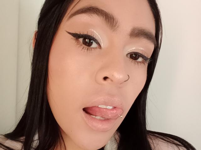 StellaBeckker's face, showing glossy pink lips, tongue out with a piercing, dark straight hair, brown eyes with glittery eyeshadow, and a nose ring, looking sexy and cute.