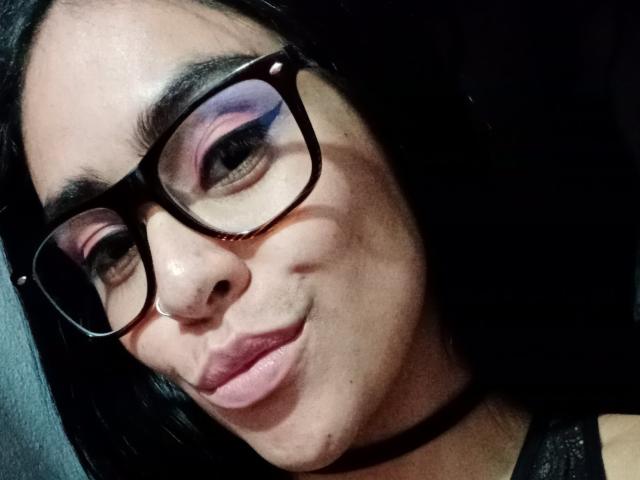 StellaBeckker with glasses, black hair, pink eyeshadow, pouty lips, and a black choker, looking slightly to the side with a subtle smirk. Her nose has a small scar.