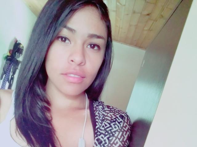 StellaBeckker with long black hair, brown eyes, pink lips, wearing a patterned top, white necklace, and a cute expression, in a bright room with a wooden ceiling.
