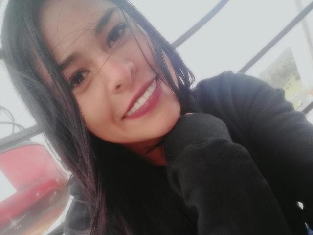 StellaBeckker with long black hair, smiling with white teeth, wearing a black top, holding her chin, looking at the camera, showing a cute face, and pink lips.