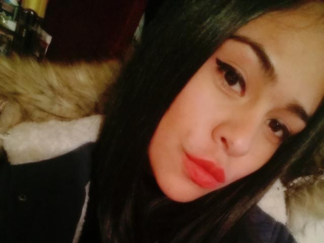 StellaBeckker has pale skin, black hair, thick eyebrows, and red lipstick. Her brown eyes look slightly to the right, and she's wearing a dark jacket with a fur trim.
