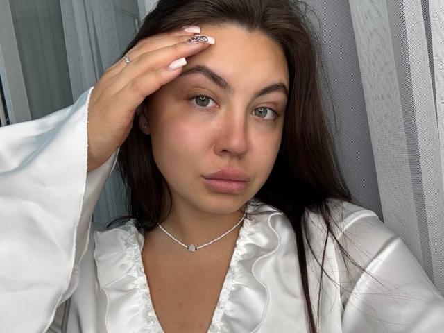 Ines Gueningsman, com cabelos castanhos longos, olhos verdes e lábios carnudos, vestindo uma camisa de cetim branca, com a mão na testa, um colar de prata e um anel, com uma expressão séria. Seios visíveis..