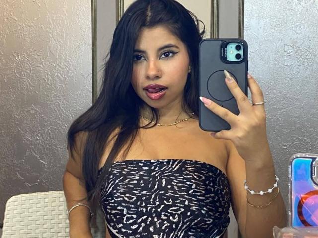 Celeste Morel, con lunghi capelli scuri, indossa un top nero e bianco senza spalline, tiene un telefono davanti al viso, con la bocca leggermente aperta, e guarda la fotocamera..