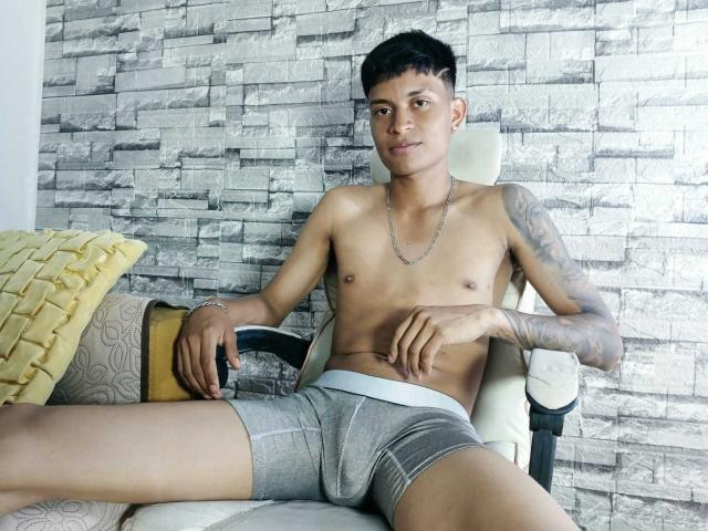 CristianDosSantos, ohne Hemd, trägt graue Boxershorts, hat tätowierte Arme, sitzt auf einem Stuhl, die Beine sind gespreizt, blickt in die Kamera, hat kurze schwarze Haare, eine silberne Kette um den Hals und gelbe und graue Kissen..