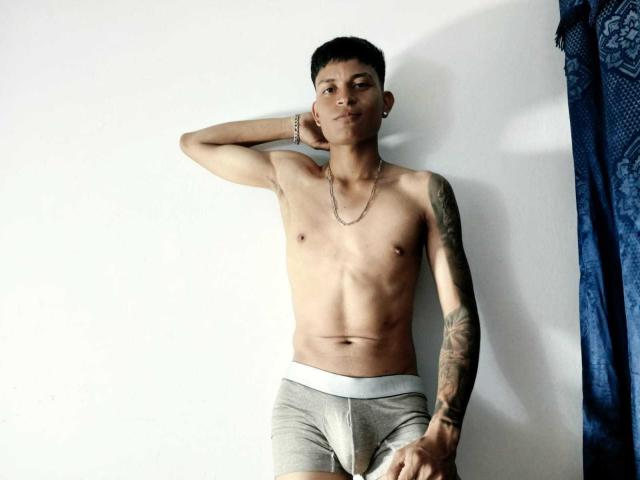 CristianDosSantos, sin camisa, con un brazo tatuado, vistiendo ropa interior gris, de pie contra una pared blanca, con la mano detrás de la cabeza, con una expresión de confianza, cabello negro corto, una ligera sonrisa, collar de cadena..