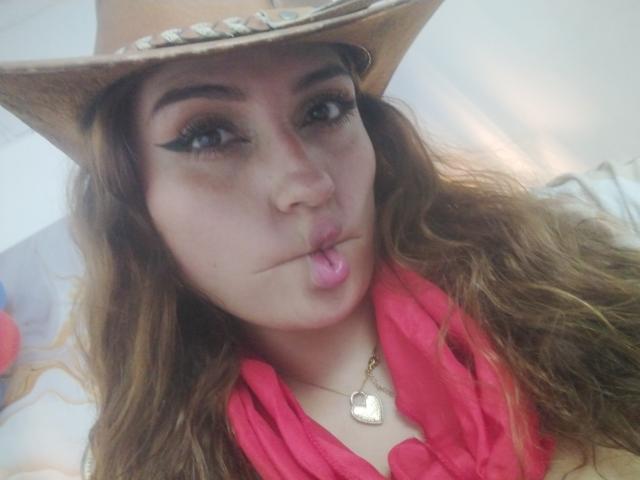 LaylaCollins with wavy brown hair, wearing a cowboy hat, black eyeliner, pink lipstick, pouting, and a pink scarf, heart necklace, brown eyes, and a cute face.