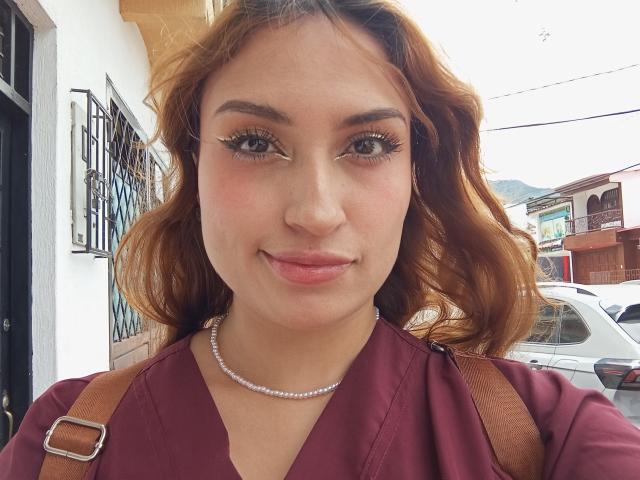 LaylaCollins with wavy brown hair, light skin, and a pearl necklace, wearing a maroon V-neck shirt, brown straps, and subtle makeup, smiling at the camera.