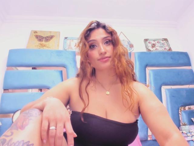 LaylaCollins with wavy brown hair, black strapless top, showing her big tits, blue upholstered chairs, tattoo on right wrist, gold necklace, and pink nails in a bright room.