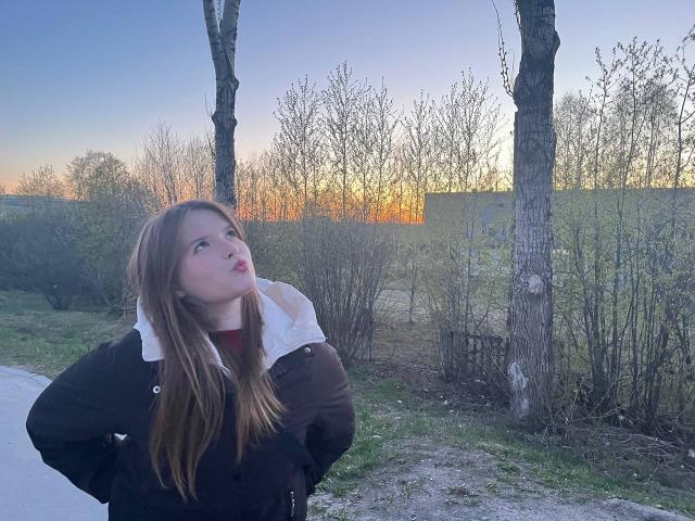 RozellaTorok has long brown hair, a white-lined black jacket, and pouts with her hands on her hips, looking up at a sunset. Two bare trees are in the background.
