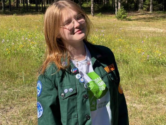 RozellaTorok with blonde hair, glasses, green scout jacket, white shirt, standing in sunny field, head tilted, eyes closed, wearing multiple colorful pins, green bag hanging.