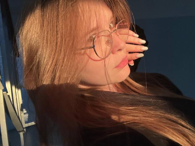 RozellaTorok with long brown hair, wearing round glasses, and pale pink lipstick, covering her mouth with her white-manicured hand, sunlight highlighting her face, in a black top.