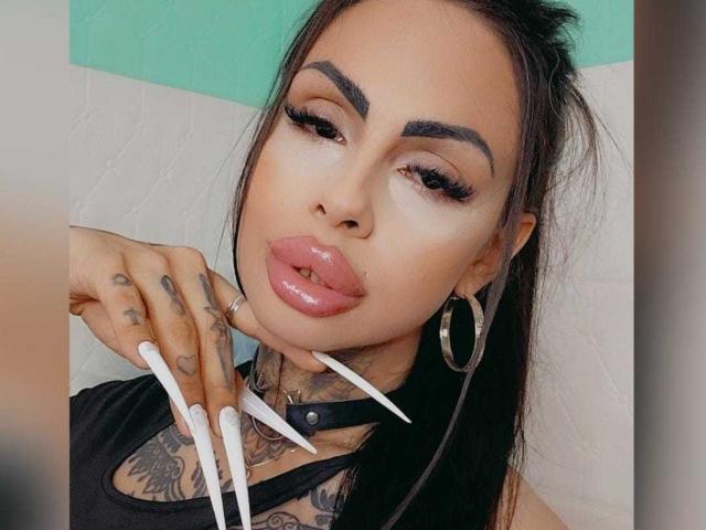 DenyChantal con unghie lunghe e bianche, tatuaggi, orecchini a cerchio, labbra rosa lucide, la lingua fuori, un collare nero e capelli scuri. Sfondo verde e bianco..