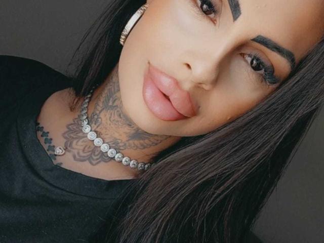 Il viso di DenyChantal, con labbra piene e carnose, sopracciglia scure, lunghi capelli neri e una collana con diamanti. Ha un tatuaggio sul collo e indossa una maglietta nera..