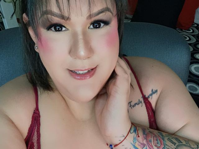 GiiannaMorrys with pink blush, black eyeliner, and nose piercing, wearing red lace top, showing tattoo on right shoulder, hand on neck, smiling, visible tattoos on right arm.