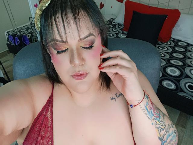 GiiannaMorrys with pink eye makeup, red lace top, nipple visible, hand touching ear, colorful tattoo on arm, red nail polish, short black hair, gold headband, slightly open mouth.