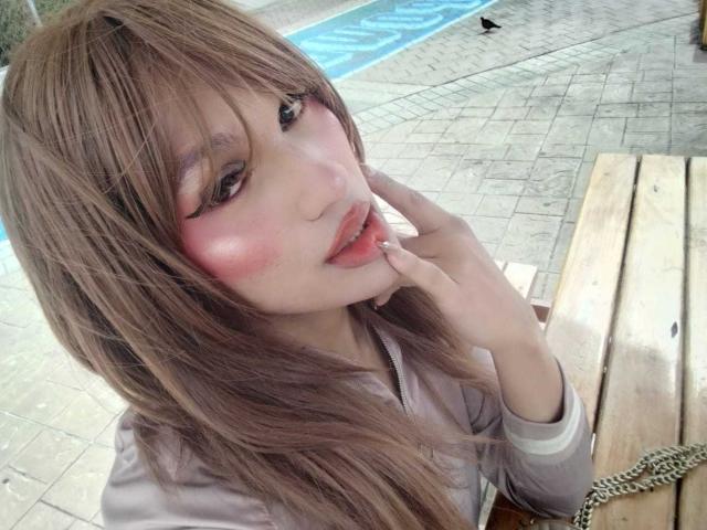 PamelaXFox with long brown hair, pink blush on cheeks, pink lipstick, and a hand near her mouth, sitting by a wooden bench, looking at the camera.