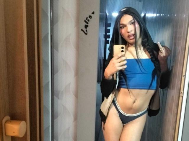 PamelaXFox met lang zwart en blond haar, een blauwe top, zwart-witte onderbroek, die een telefoon vasthoudt, een spiegelzelfportret, met een lichte grijns, draagt een zwarte en beige jas, met een getoonde taille..