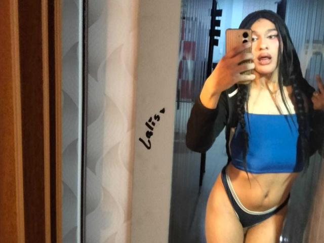 PamelaXFox with long black hair, blue crop top, black panties, holding phone in mirror selfie, showing her tits and pussy, with a tattoo of "Uliis" on the mirror.