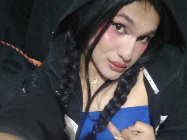 PamelaXFox with braided black hair, pink eye makeup, and red lipstick, wearing a black hoodie over a blue top, pointing at her chest with a ring on her finger.