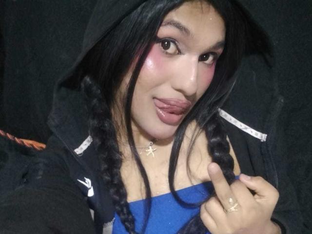 PamelaXFox with black braids, pink blush, and tongue out, wearing a black hoodie, blue top, and cross necklace, holding up a finger, looking at the camera.