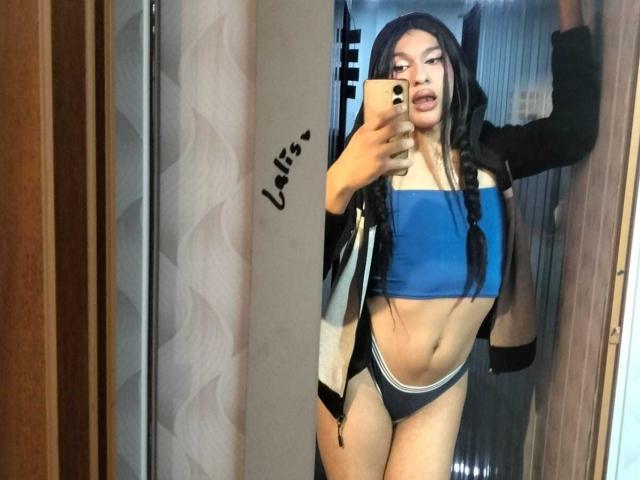 PamelaXFox, avec de longs cheveux noirs, portant un débardeur de sport bleu et des sous-vêtements noirs, se tient devant un miroir et prend un selfie avec son téléphone. Ses seins sont petits..
