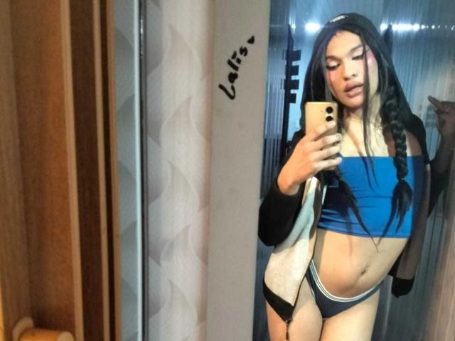 PamelaXFox con lunghi capelli neri, reggiseno sportivo blu, biancheria intima nera e bianca, che tiene il telefono per un selfie allo specchio, mostrando piccoli seni e un sedere snello e tonico, indossa una giacca nera e bianca..