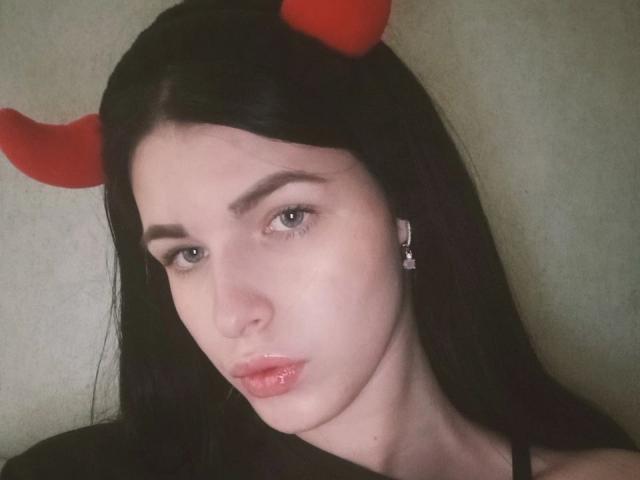 Dianne Zarrineh tem pele clara, cabelo preto e chifres vermelhos de diabo. Ela está usando um batom rosa brilhante e tem uma expressão séria, com os lábios levemente entreabertos e rosados..