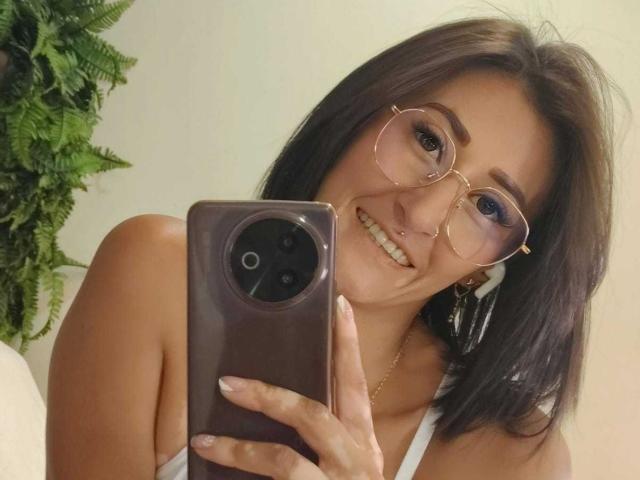 SharissWilliam with brown hair, wearing glasses, smiling, holding a phone, showing off her tits in a white top, and a nose ring. Green plant on the left.