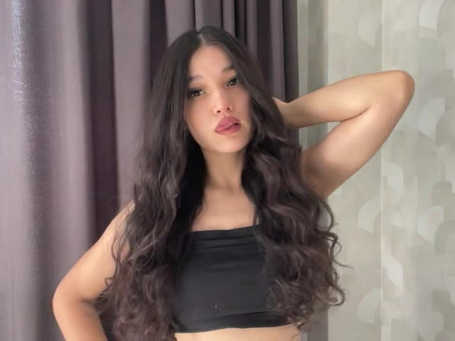 YoulandaCarlough with long, wavy black hair, wearing a black crop top, standing with one arm behind her head, looking at the camera with pouty red lips.