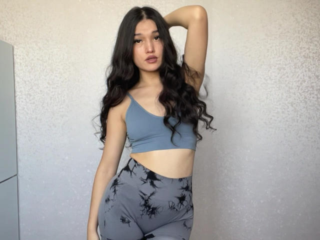 YoulandaCarlough with long black wavy hair, gray sports bra, and gray high-waisted leggings with black bird patterns, arm raised, showing tattoo on left arm.
