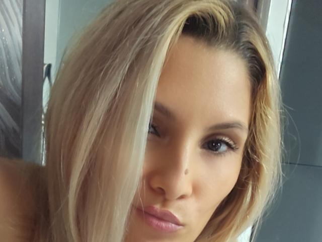 DianeExo has blonde hair, winking, and a cute mole on her nose. Her pink lips are slightly pursed. She's looking at the camera with one eye open.