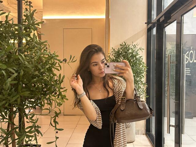 EmeraldCrivaro with long brown hair, pouty lips, black dress, striped cardigan, brown bag, taking mirror selfie, holding phone, standing near green plant, warm lighting, inside.