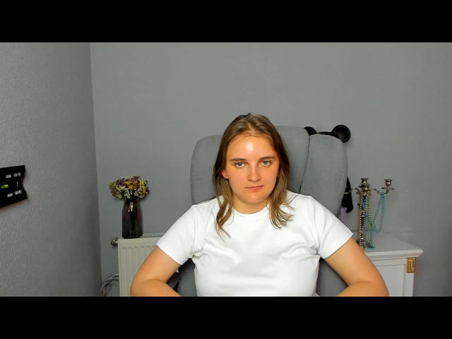 Bileira, with blonde hair and blue eyes, sits in a grey chair, wearing a white shirt, looking straight at the camera with a neutral expression.
