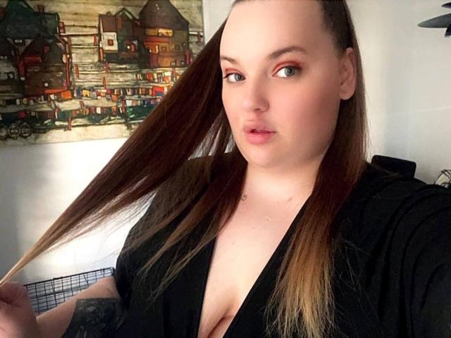 DevilishMuse heeft bleke huid, lang bruin haar en houdt een haarstreng vast. Ze draagt een zwarte top met een diepe V-hals, waardoor haar kleine borsten zichtbaar zijn..