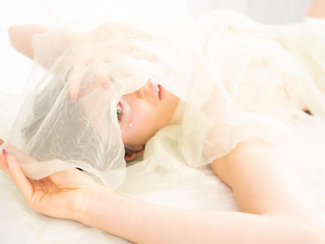 Sarah está acostada en una cama blanca, semidesnuda, con un tejido blanco translúcido que le cubre la cabeza y el torso superior, mostrando sus pechos y un atisbo de su vulva. Iluminación suave y etérea..