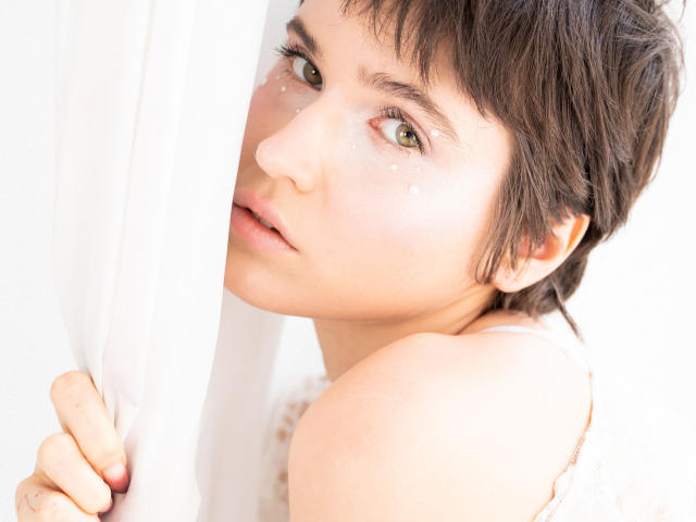 SarahMiss ha i capelli castano corti, gli occhi verdi e dei piccoli puntini bianchi scintillanti sulla guancia. Tiene in mano una tenda bianca, guarda leggermente a sinistra con un'espressione dolce..