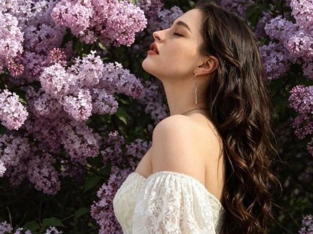DorianCieslinski with eyes closed, head tilted back, long wavy brown hair, white lace off-shoulder top, small tits, surrounded by pink lilac blossoms, wearing simple gold earrings.