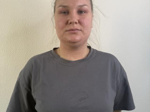 ValerieSchultheiss has a fat face, light skin, and small tits, wearing a gray shirt, with her hair tied back, standing against a white textured wall.