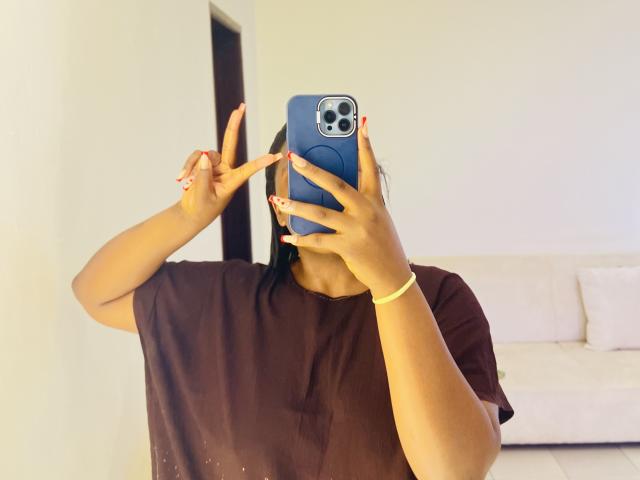 ThickMari with tan skin, black hair, brown tee, holding blue phone, flashing peace sign, yellow bracelet, red nails, taking selfie in bright room.